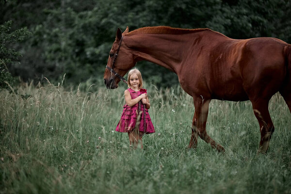 Beautiful little blonde girl in a dress next to a big red horse