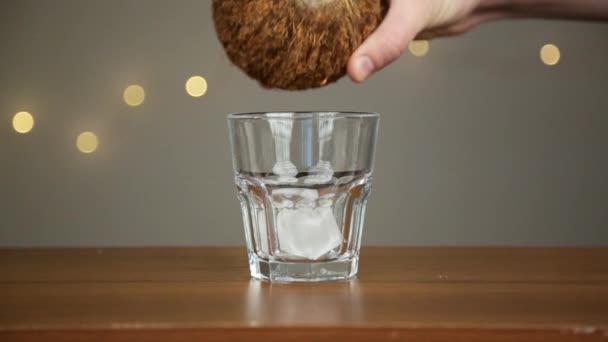 Man kastar is i ett glas, sedan häller kokos vatten. Mot bakgrund av lamporna — Stockvideo