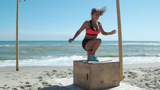 Blond femme trains jambes saute sur la boîte Crossfit avec un entraîneur Sur la plage — Video