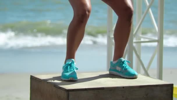 Mujer rubia entrena piernas Salta a la caja Crossfit con un entrenador En la playa De cerca — Vídeo de stock