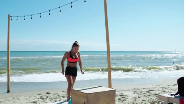 Mujer rubia entrena piernas Salta a la caja Crossfit con un entrenador En la playa — Vídeo de stock