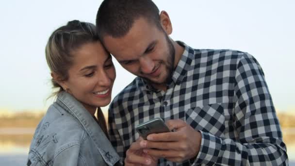 Strandabend Ein paar junge Verliebte blicken in ein Smartphone und lächeln — Stockvideo