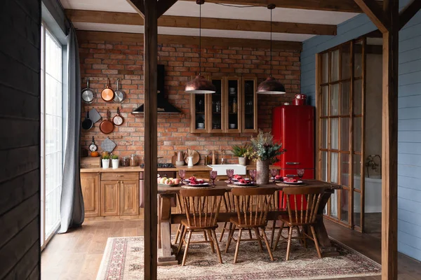 カントリーハウスのキッチン 木製のキッチンには 大きなテーブルと椅子が床にパノラマの窓の近くに立って 赤い冷蔵庫 青い木製の壁 レンガの壁にぶら下がっているパンが置かれています ロフトスタイル — ストック写真