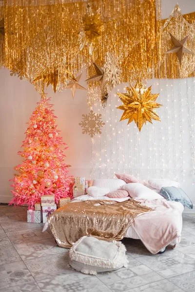 Christmas morning interior with pink christmas tree decorated bed and big window. White, pink and gold decorations. New Year. Beige armchair. A lot of pillows. Golden Christmas star. Luxury interior.