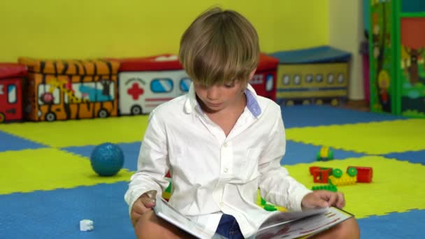 Słodki chłopiec czyta książkę na podłodze w pokoju zabaw. — Wideo stockowe