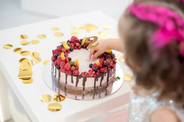 birthday cake with fruits and nuts on a light background with confetti, with the number 2 and the baby from the back