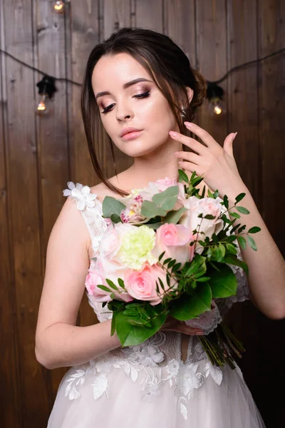 Slim beautiful woman wearing luxurious wedding dress on wooden background. Gorgeous bride holding flowers. — Stock Photo, Image