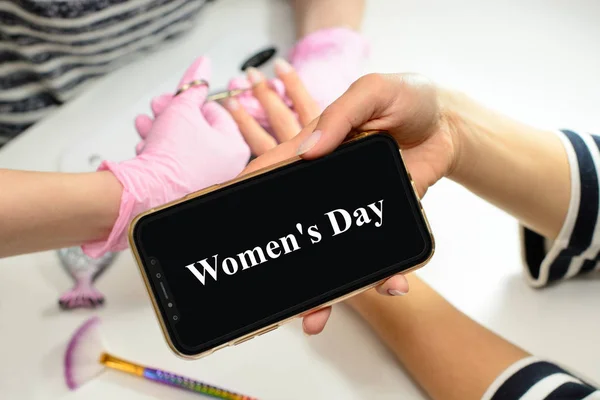 Nő használja a mobiltelefon, a telefonon a felirat Women Day, nő szalonban kap egy manikűr és körömápolási eljárás kozmetikus. Nő egyre körmök által végzett manikűrös szépségszalonban. — Stock Fotó