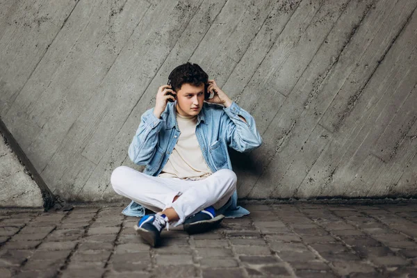 Portrait of handsome young man with denim shirt sits on a concrete floor and listens to music with black headphones