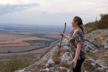 Yaşlı bir kadın bir dağın tepesinde duruyor, bir sopaya yaslanıyor ve gülümsüyor.