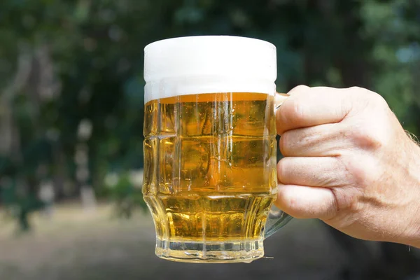 glass mug with beer and foam in a man\'s hand on the background of nature close up