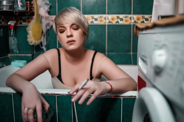 Young blonde woman in the bathroom sitting drinking whiskey and — ストック写真