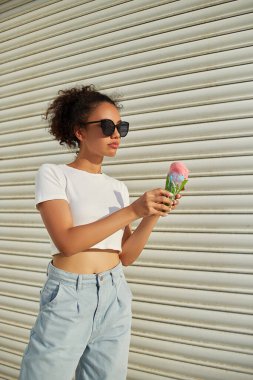 Beyaz tişörtlü ve açık kotlu güzel bir Afro-Amerikalı kız güneşli bir günde açık renkli bir duvarda dondurma yiyor..