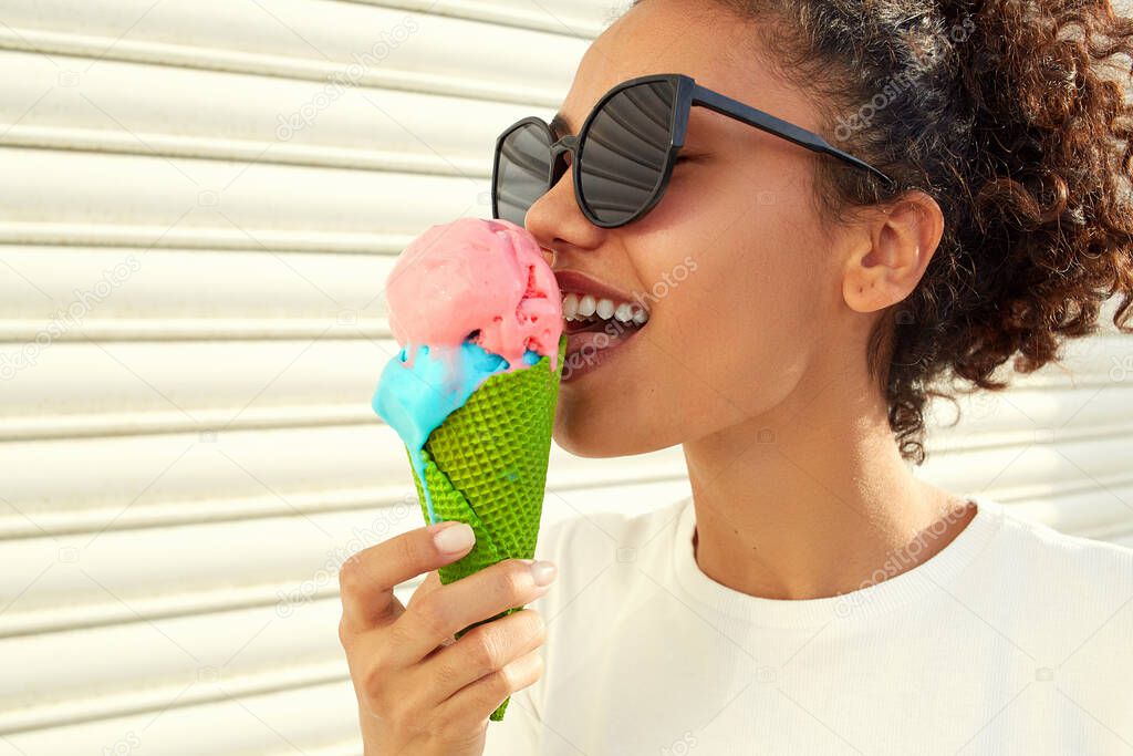 a young beautiful African-American girl in a white t-shirt and light jeans eats ice cream against a light wall on a Sunny day.
