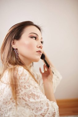 Bej dantelli sabahlık giyen, sabah makyajı yapan ve göz farı, allık ve fondöten kullanan güzel bir Asyalı kadın. Yatak odasında. Sığ odak derinliği. seçici odak.