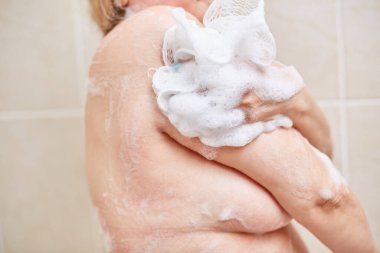 60 yaşında yaşlı bir kadın duşta yıkanıyor, omuzlarını, sırtını ve vücudunu banyo beziyle sabunluyor. seçici odak, küçük odak alanı