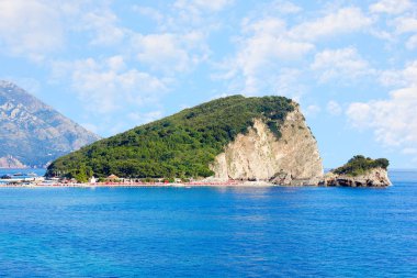 Karadağ'daki Budva yakınındaki St. Nicholas Adası. Mavi deniz, kayalar ve gökyüzü bulutlu sahil.