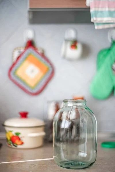 Empty three-liter jar is on the tabletop against the background of the pot and oven mitts. — Stock Photo, Image