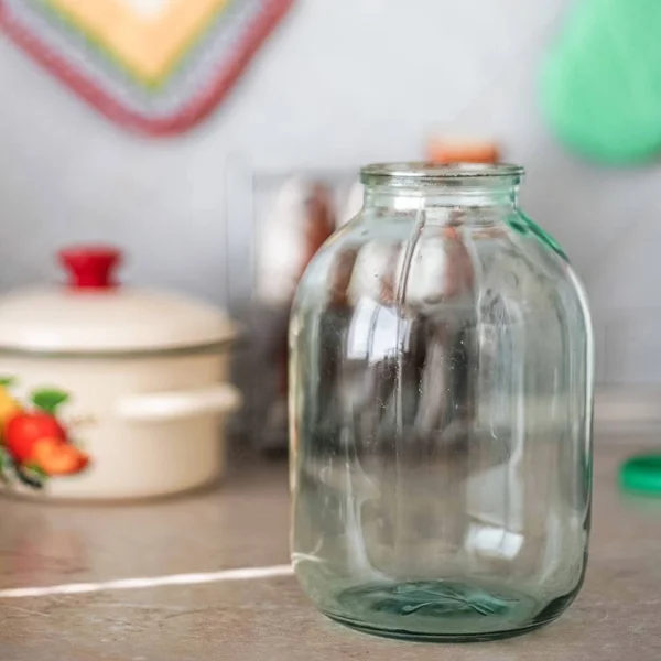 Empty three-liter jar is on the tabletop against the background of the pot and oven mitts. — Stock Photo, Image