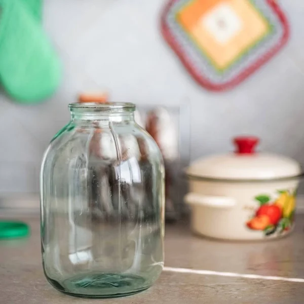 Empty three-liter jar is on the tabletop against the background of the pot and oven mitts. — Stock Photo, Image