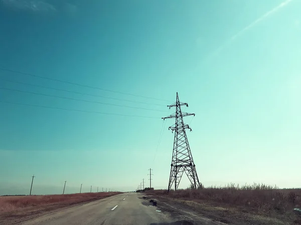 Asphaltstraße durch das Feld unter den elektrischen Leitungen. Stromleitungen führen durch braune Felder. — Stockfoto