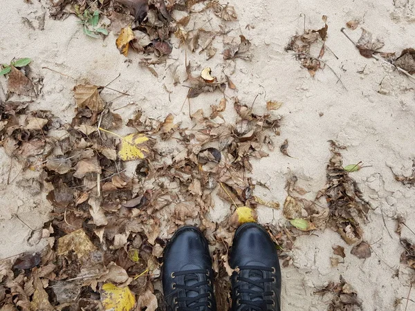 Bottes noires sur une plage de sable parmi des feuilles d'automne brunes et sèches . — Photo