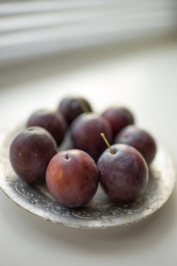 Gümüş bir tabakta olgun mor erikler. Beyaz masada taze meyveler. Yan görünüm