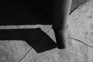 Binanın köşesindeki borudan. Taş zeminde çatlaklar var. Parlak güneş ışığı ve kanalizasyon borusundan gelen gölge. Siyah beyaz fotoğraf.