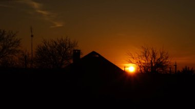 Karanlık günbatımında bir köy evinin ve ağaçların siyah silueti. Turuncu gökyüzünde yuvarlak güneş