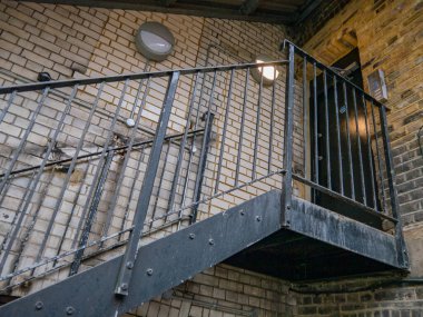 Tuğla bir duvarda acil çıkış kapısı. Caddeye çıkan metal merdivenler var. Yumuşak sarı bir ışık veren küçük bir lamba var..