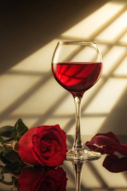 Bir bardak kırmızı şarap ve masada yansımalı kırmızı bir gül, pencereden gelen ışık.