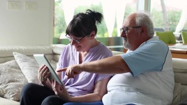 Yaşlı bir adam, kadınlara tablet kullanmayı öğretirken bir koltukta oturuyor, gülümsüyor ve modern bir oturma odasında dijital öğrenmenin tadını çıkarıyor..