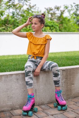 11 yaşında, renkli paten ve günlük kıyafetlerle beton bir çıkıntıda güneşli bir günün tadını çıkaran bir kız..