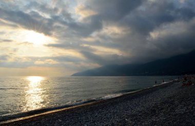 Dağların yakınındaki deniz çakıl plajı üzerinde dramatik kabarık bulutlar. Güneş bulutların arasından parlar ve suya yansır. Dalga kıyıya doğru koşuyor.