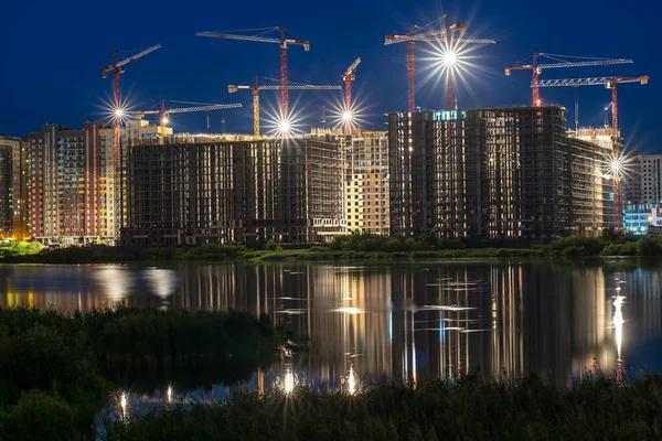 multistory buildings under construction made of reinforced concrete structures and frameworks with cranes at night near pond with reflected lights and glare in water.