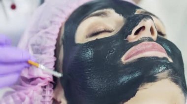 Mor eldiven makyajcısı dikkatle siyah maske arındırıcı yüz genç bir kadın koyar. Scin bakım, güzellik uzmanı yüz maskesi krem uygulamak.