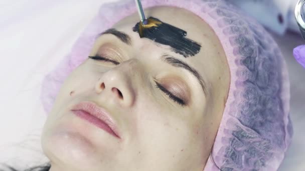 Cosmetologist esfrega máscara cosmética no rosto da mulher no salão de spa. Mulher bonita em boné médico recebendo máscara preta no rosto. Close-up — Vídeo de Stock