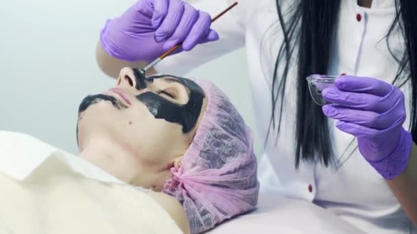 美しい若い女性は、スパで寝ながら顔のマスクをことです。女性は美容師によって泥黒マスク化粧品の手順を取得します。. — ストック動画