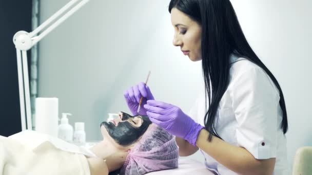 Atractiva esteticista en abrigo médico y guantes azules aplicando máscara cosmética negra en la cara a una mujer en el salón de belleza. Salud y cuidado de la piel . — Vídeos de Stock