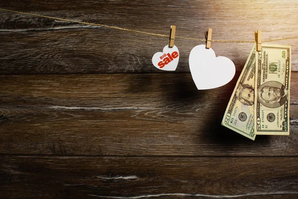 Two hearts and dollar banknotes drying on the clothesline by clothespins on wooden background. Spring sale