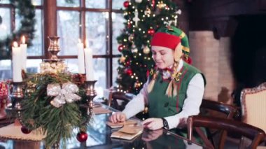 Cheerful elf girl sitting at a wooden table looking through Santa Claus mail. Room with Christmas decorations.