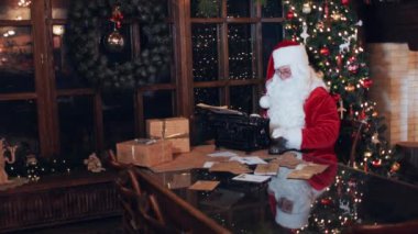 Santa Claus at a table near a Christmas tree prints a letter to children
