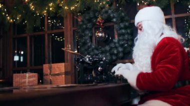Santa Claus sits at a table at a typewriter in an evening room with New Year decorations and gifts.