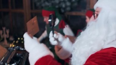 Santa Claus at the table looks through letters through a magnifying glass. Elf is played on the background.