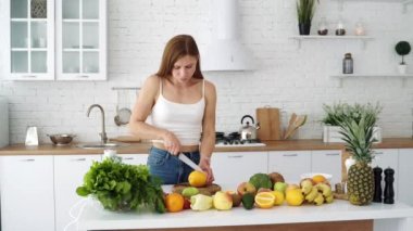 Bir kadın taze bir portakalı bıçakla keser ve koklar. Vejetaryen yemeği. Sebze ve meyveli beyaz modern mutfak..