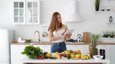 Elinde tablet olan bir kız sağlıklı yemek pişirmek için tarifler arıyor. Masada meyve ve sebzelerle dolu beyaz bir mutfak..
