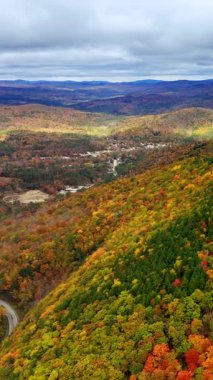Dağların arasındaki bir vadideki köyün havadan görünüşü. Yamaçlarda renkli ağaçlarla dolu sonbahar manzarası. Mavi bulutlu gökyüzü. Dikey video.