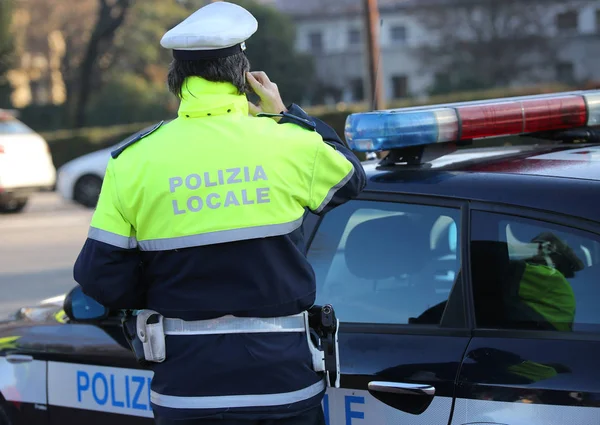 Italian policeman with the  text POLIZIA LOCALE that means local police in Italian language uses the cell phone during an emergency call and the police car