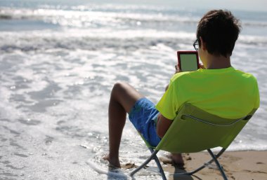 Genç Genç deniz kenarında bir katlanır sandalye üzerinde bir dijital kitap okur