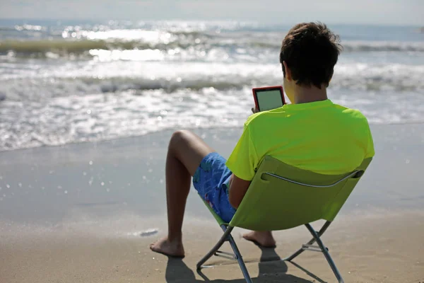 Nastolatek Czyta Książkę Cyfrowych Siedząc Krzesło Naprzeciwko Morza — Zdjęcie stockowe